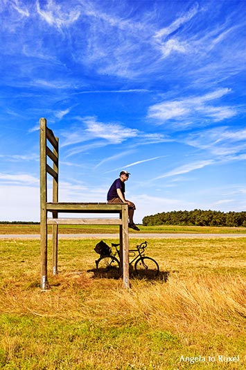 Man sitting on a chair with his bike below, oversized chair, animal shed on a field near Born, side view - Fischland/Darß 