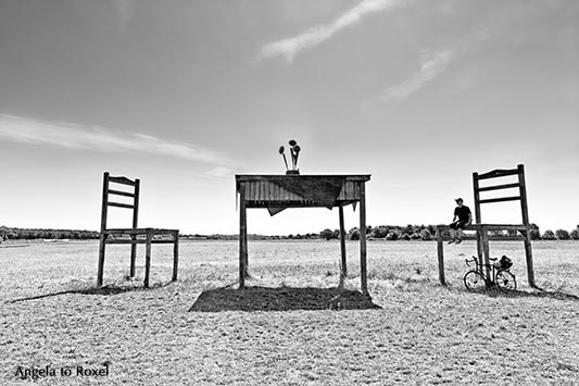 Man sitting on a chair with his bike below, oversized table and chair, animal shed on a field near Born, black and white - Fischland/Darß