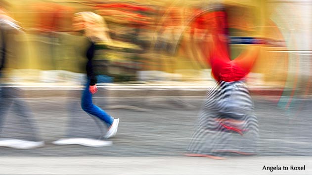 Zwei Passanten gehen in der Bielefelder Innenstadt an Schaufenstern vorbei, ein Clown schlägt hinter ihnen ein Rad, Bewegungsunschärfe - Bielefeld